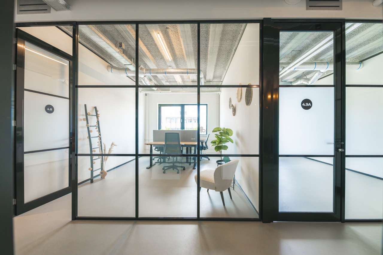 Modern ingericht kantoortje met glazen wanden, zitplekken, planten en decoratie, gezien van buiten de ruimte.