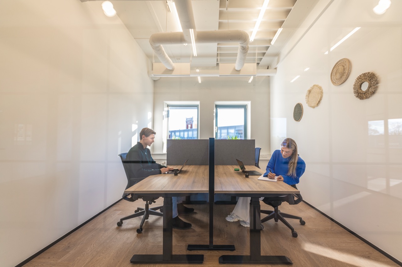 Twee mensen werken tegenover elkaar in een moderne, lichte kantoorruimte met houten vloer en wanddecoratie.