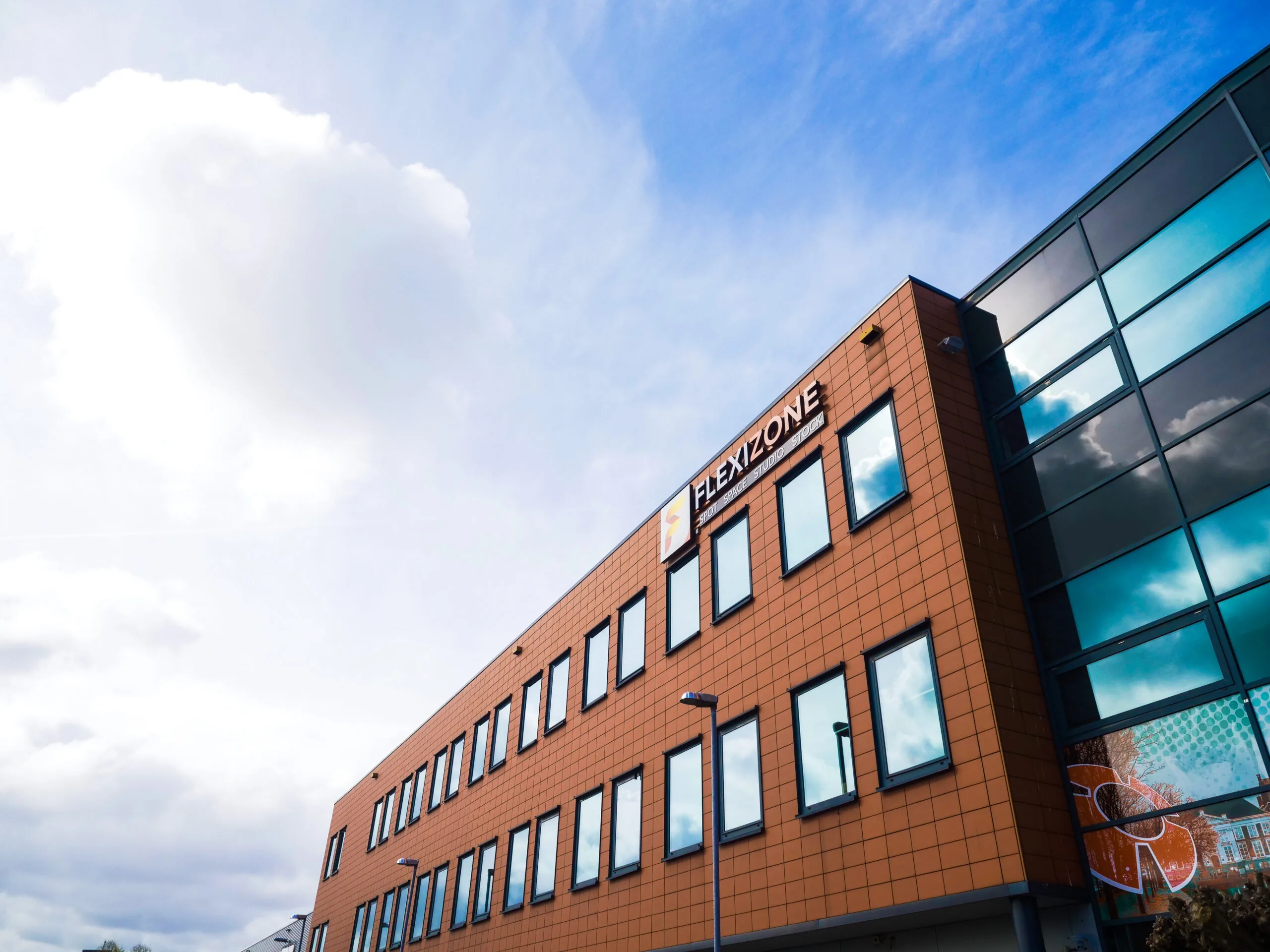Bedrijfspand met oranje gevel en het logo van FLEXIZONE onder een deels bewolkte lucht.