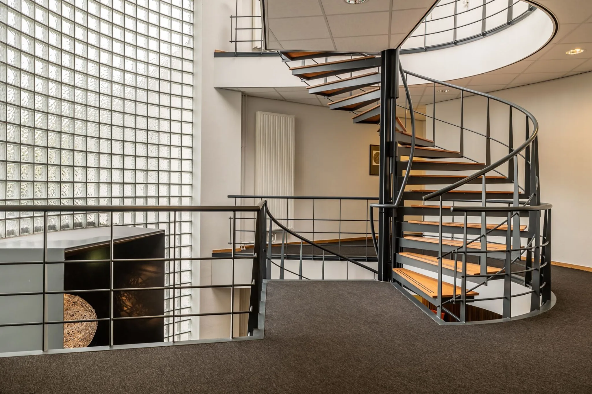 Moderne wenteltrap met metalen leuning en houten treden in een ruim en licht trappenhuis met glazen bouwstenenwand.