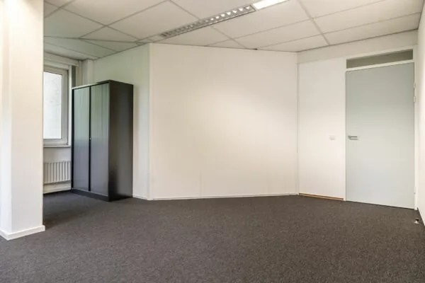 Leeg kantoor met grijze vloerbedekking, witte muren, een grijze deur en een zwarte archiefkast bij het raam, in Paardeweide 2A-4C.