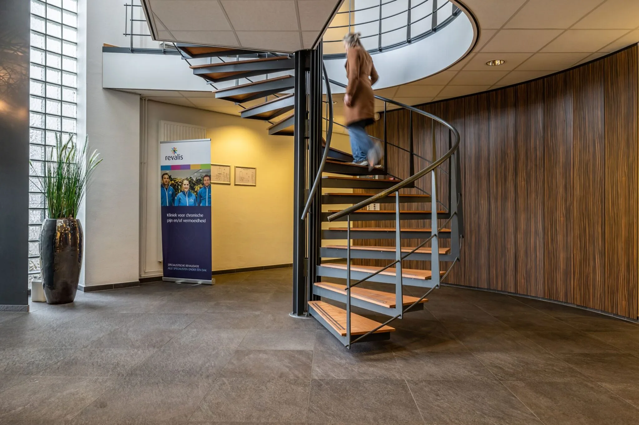Vrouw loopt een moderne wenteltrap op in de entreehal van Revalis, kliniek voor chronische pijn en vermoeidheid.