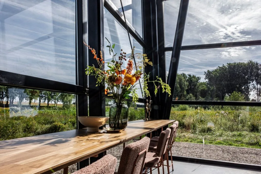 Lichte eetruimte met houten tafel, bloemenvaas en uitzicht op een groen landschap bij Plaatweg 15.