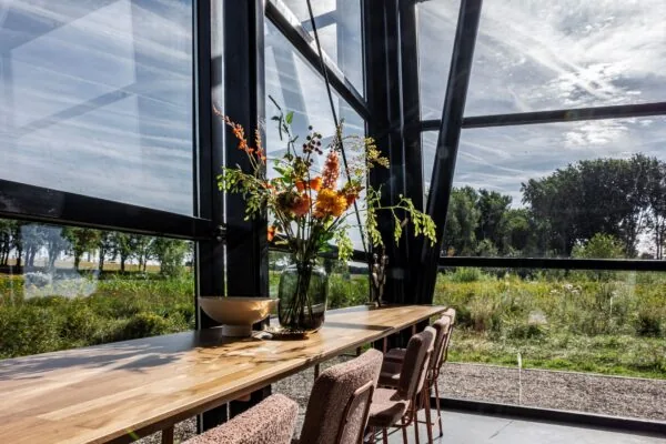 Lichte eetruimte met houten tafel, bloemenvaas en uitzicht op een groen landschap bij Plaatweg 15.