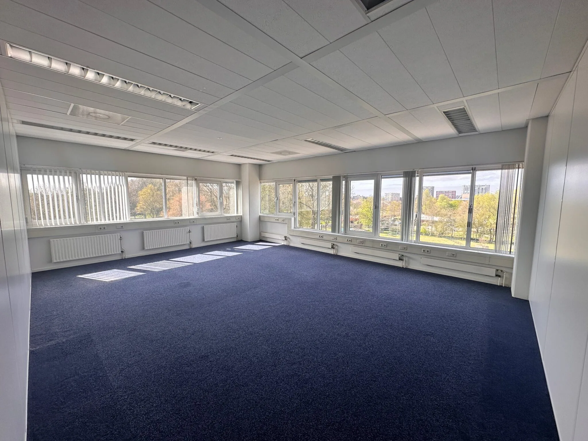 Lichte, lege kantoorruimte met blauwe vloerbedekking en grote ramen aan twee zijden, gelegen aan Planetenweg 5.