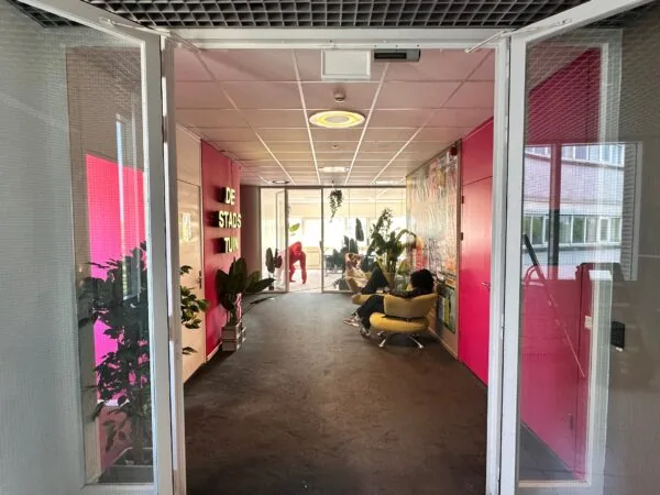 Ingang van een kleurrijke gang bij De Stadstuin aan Planetenweg 5 met planten, zitplekken en mensen die ontspannen.