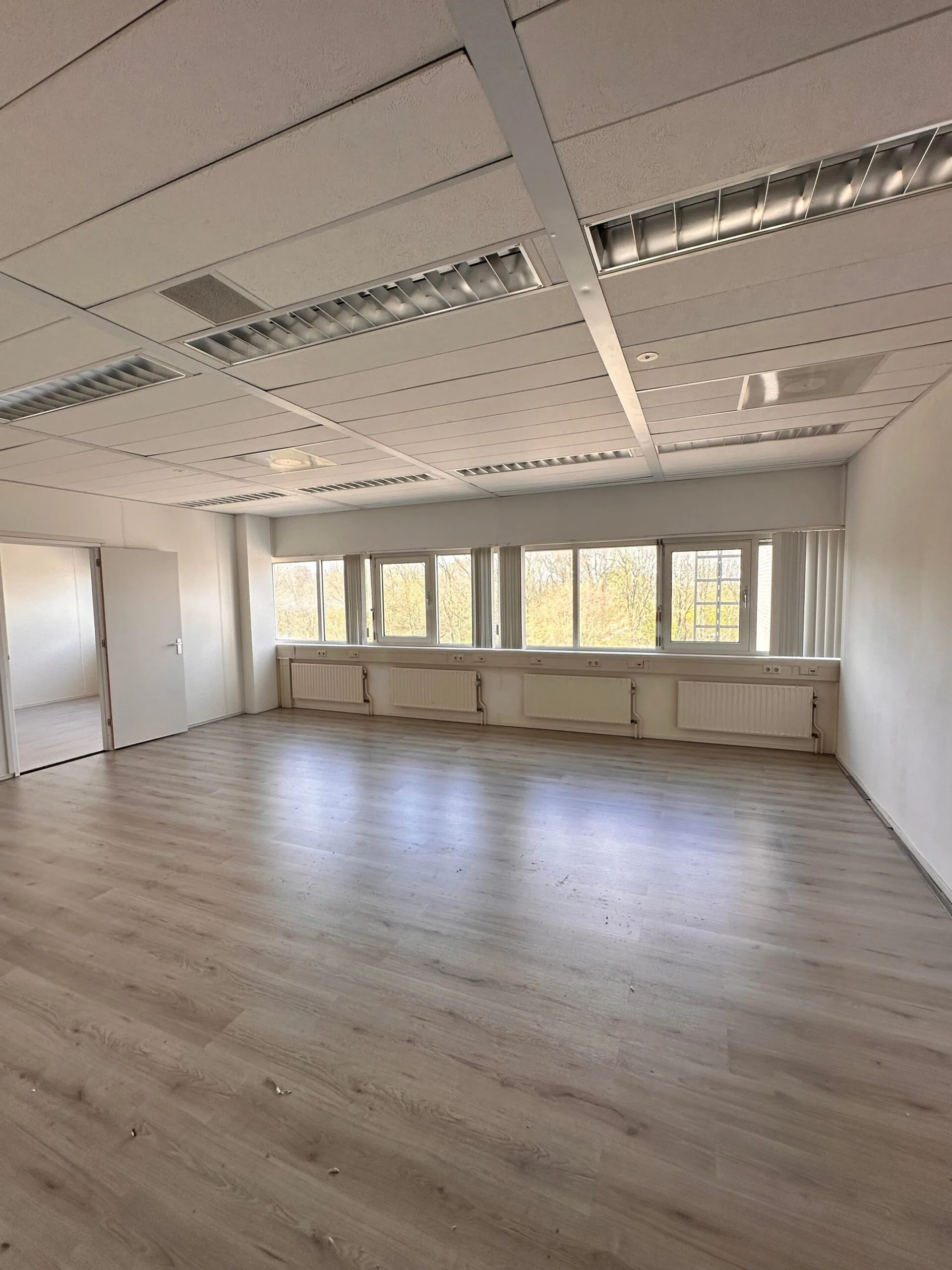 Lichte, lege kantoorruimte aan de Planetenweg 5 met grote ramen en laminaatvloer.