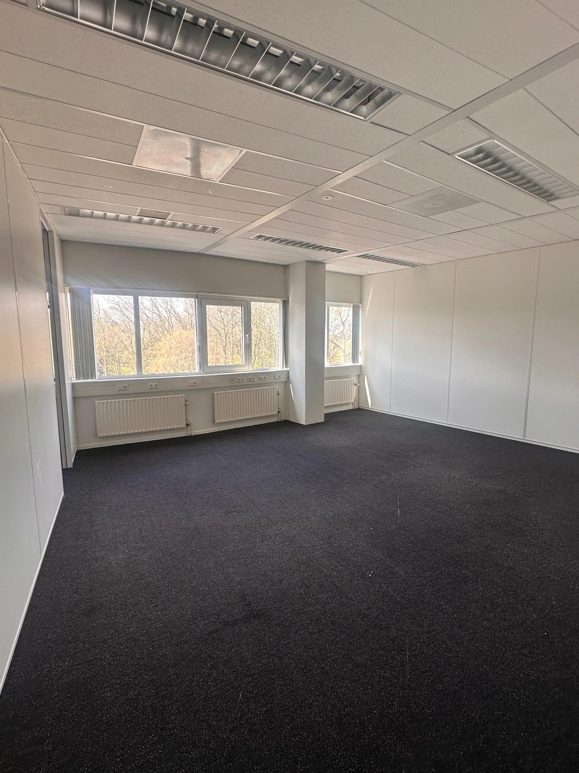 Leeg kantoor met donker tapijt, witte wanden en grote ramen bij Planetenweg 5.
