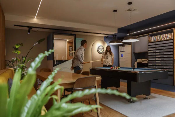 Twee mannen spelen een potje pool in een modern ingerichte recreatieruimte met planten, boeken en een dartbord aan de muur.