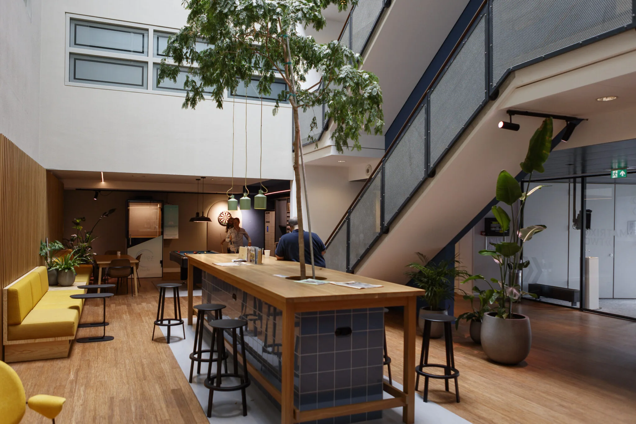 Moderne, open kantoorlounge met zithoeken, een hoge tafel, planten en een trap naar de bovenverdieping.