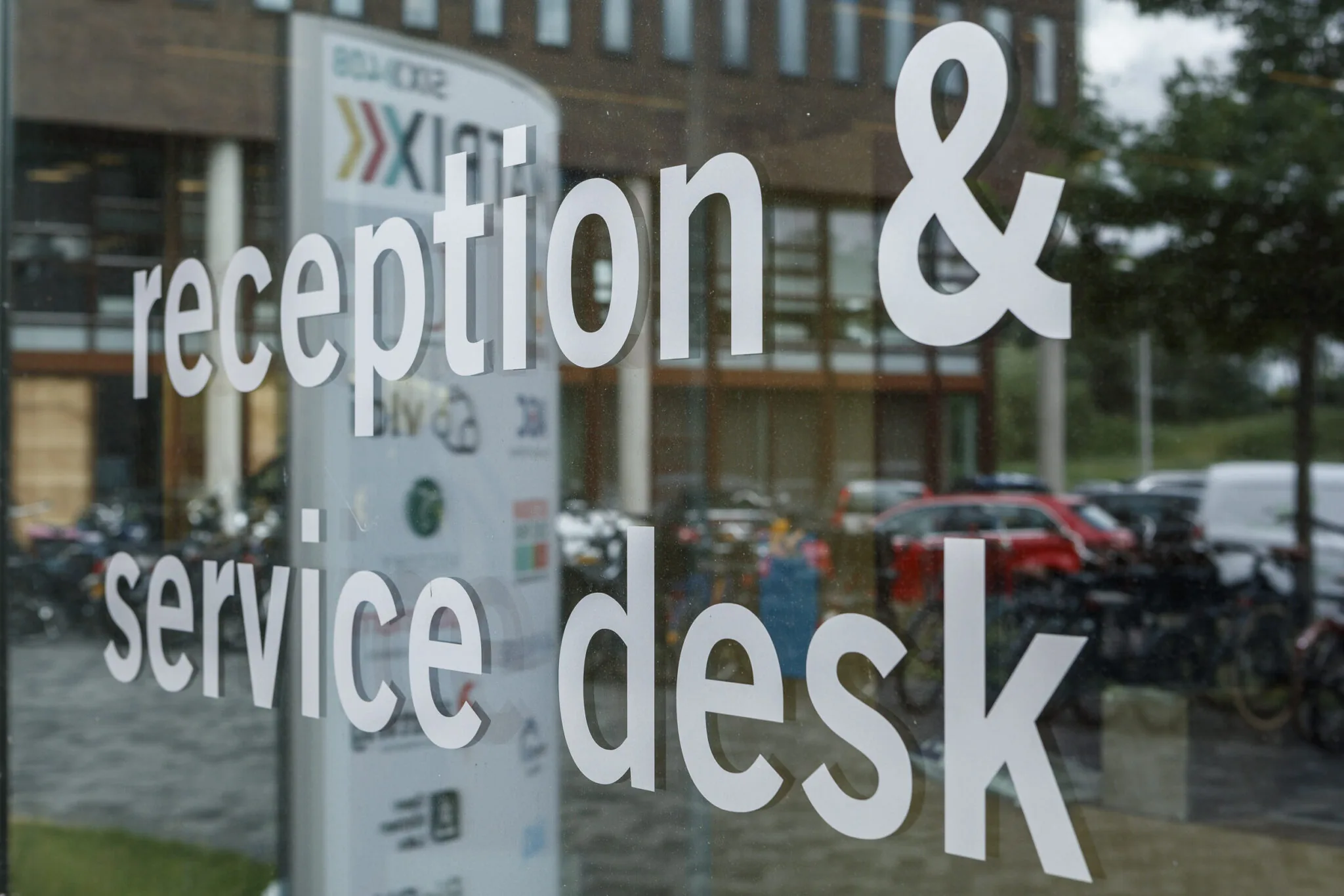 Witte tekst met "reception & service desk" op een glazen deur met reflectie van auto's en een gebouw op de achtergrond.
