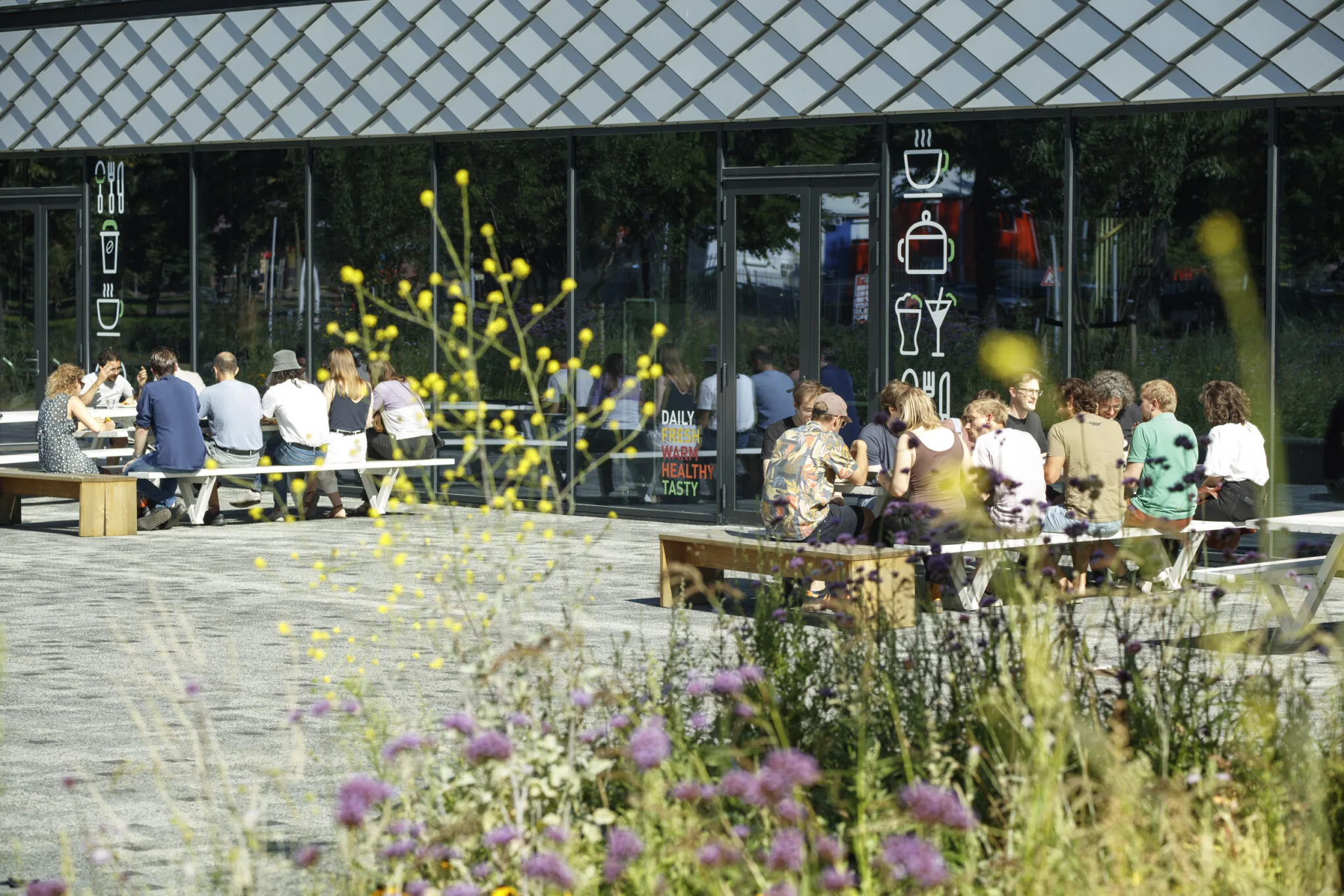 Groep mensen zit buiten op banken te lunchen bij een modern horecapand, omringd door wilde bloemen.