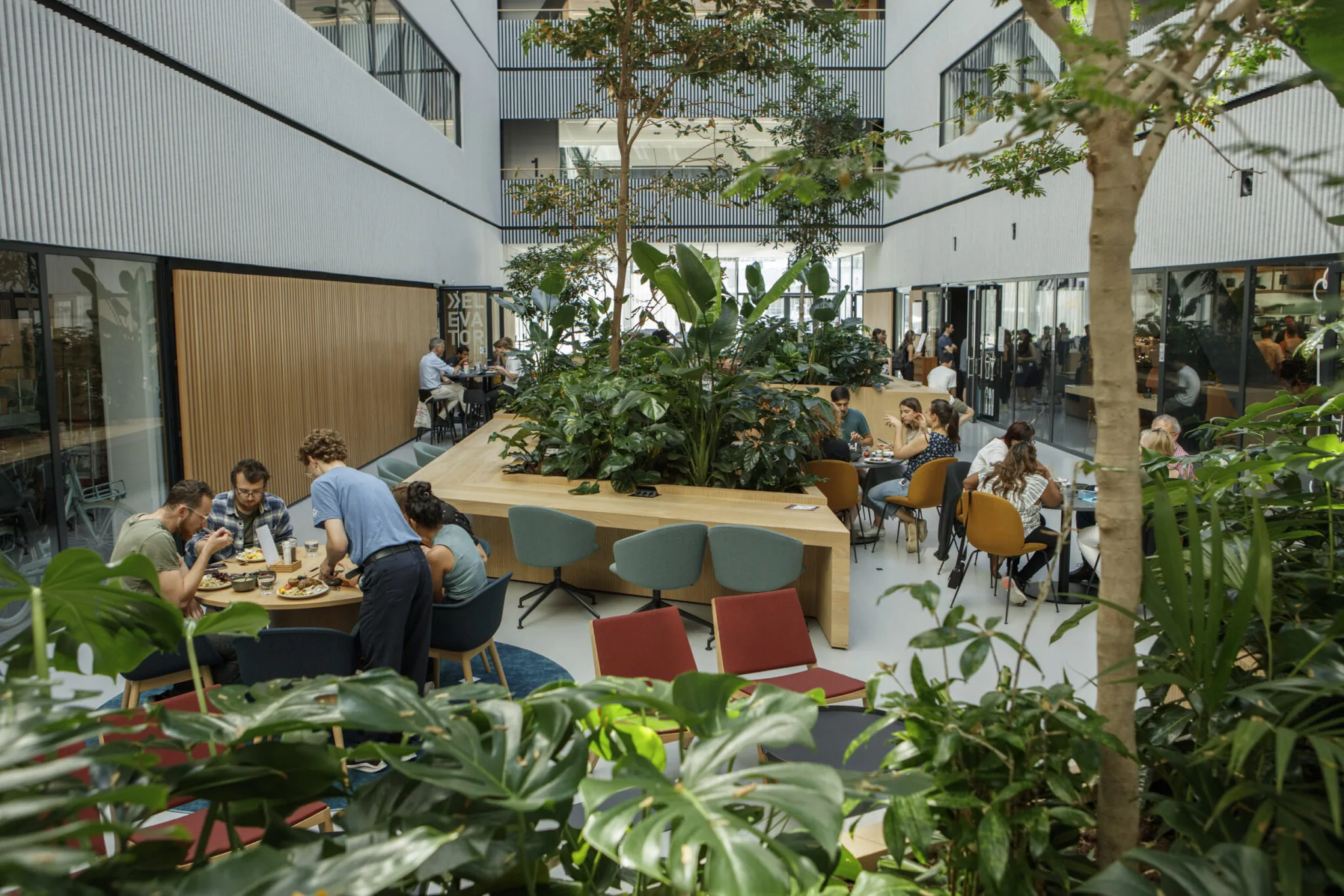 Mensen lunchen en werken in een modern, groen ingericht atrium met veel planten en natuurlijk licht.