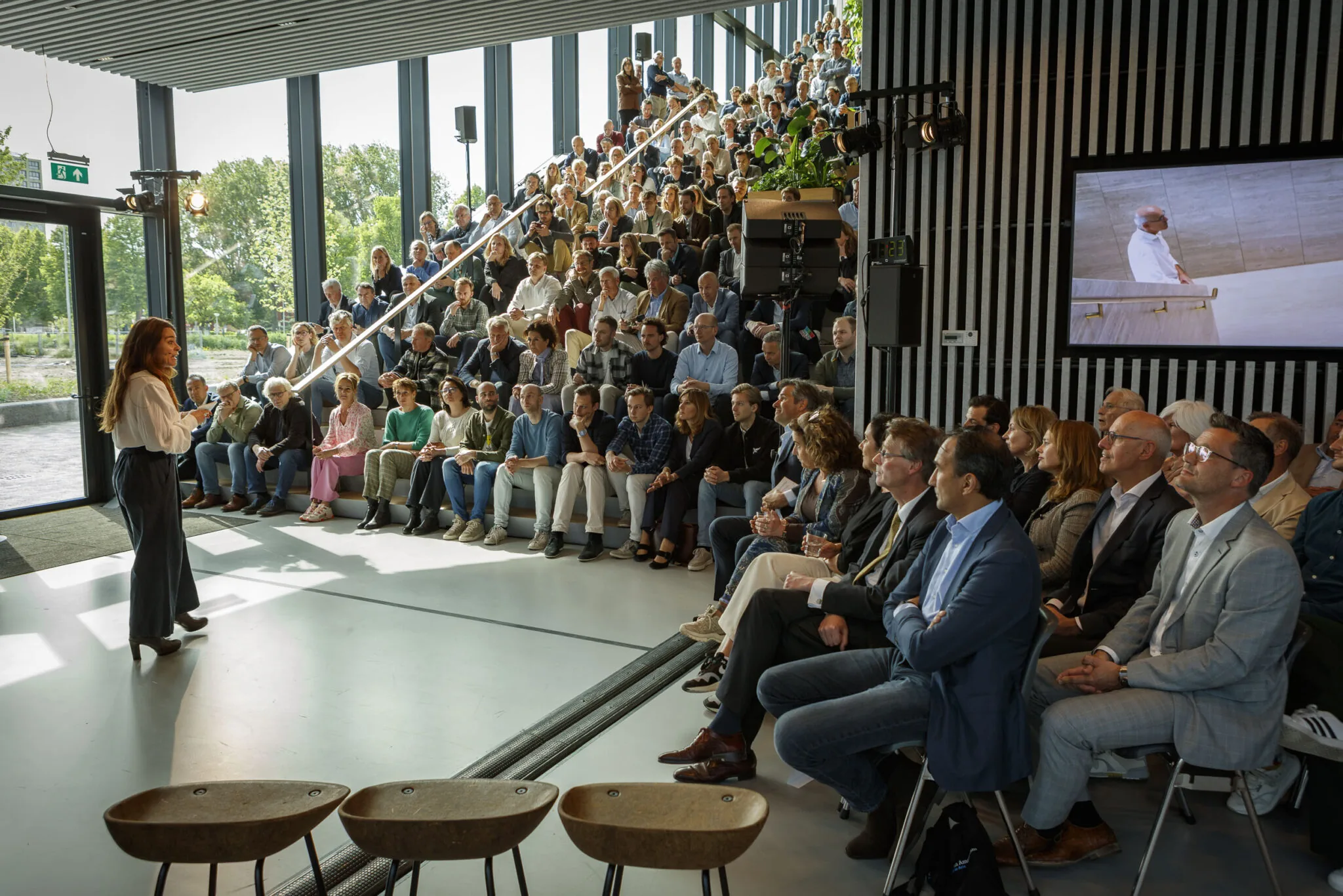 Een spreker presenteert voor een groot, aandachtig publiek in een moderne, goed verlichte ruimte met tribune.