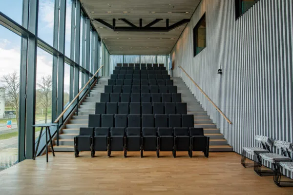 Moderne collegezaal met zwarte stoelen in een open ruimte met hoge ramen en houten vloer.