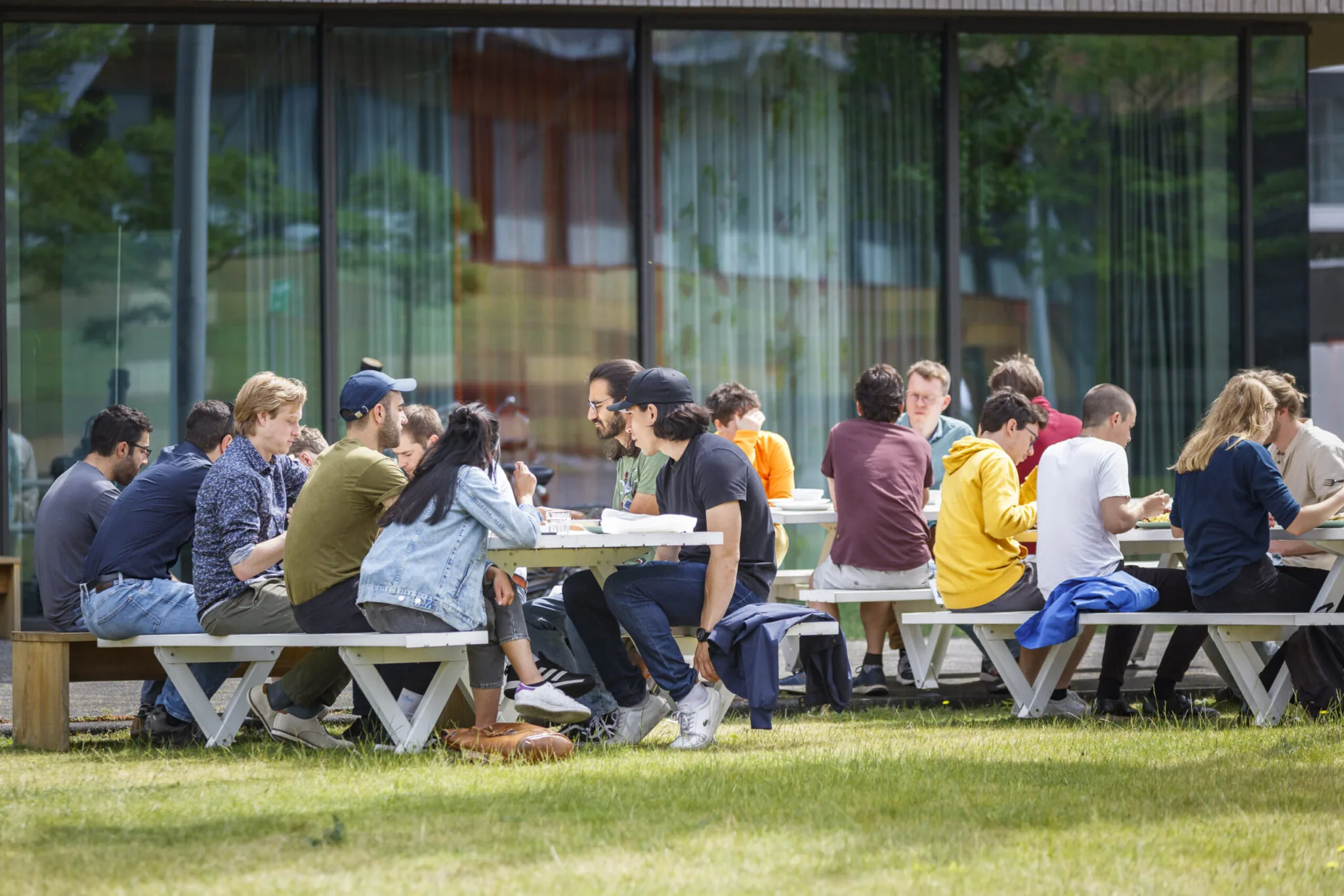 Een groep mensen zit buiten aan picknicktafels te lunchen en praten met elkaar.
