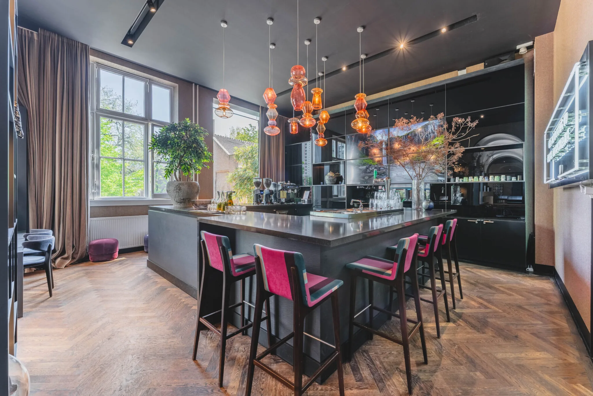 Moderne bar met roze barkrukken, een grote plantenpot en opvallende glazen hanglampen in een stijlvol interieur aan het Raamplein.