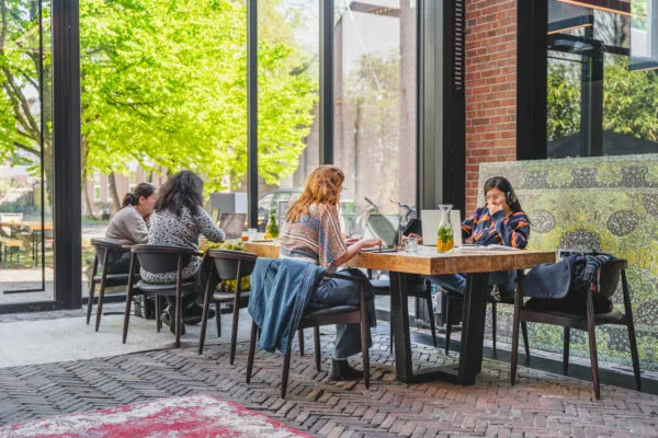 Mensen werken op laptops aan houten tafels bij Raamplein 1 in een licht en modern café met uitzicht op een groene buitenomgeving.