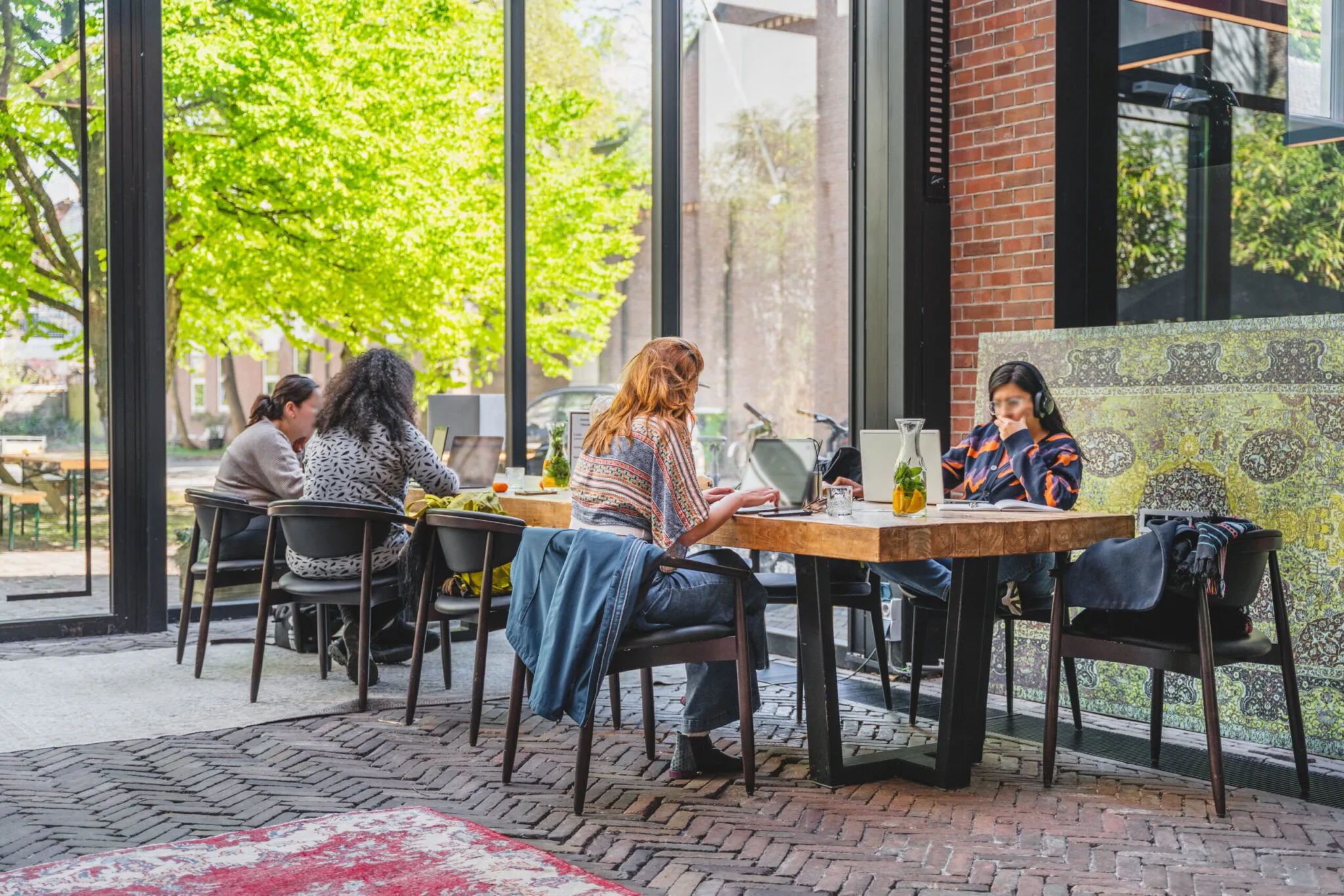 Mensen werken op laptops aan houten tafels bij Raamplein 1 in een licht en modern café met uitzicht op een groene buitenomgeving.