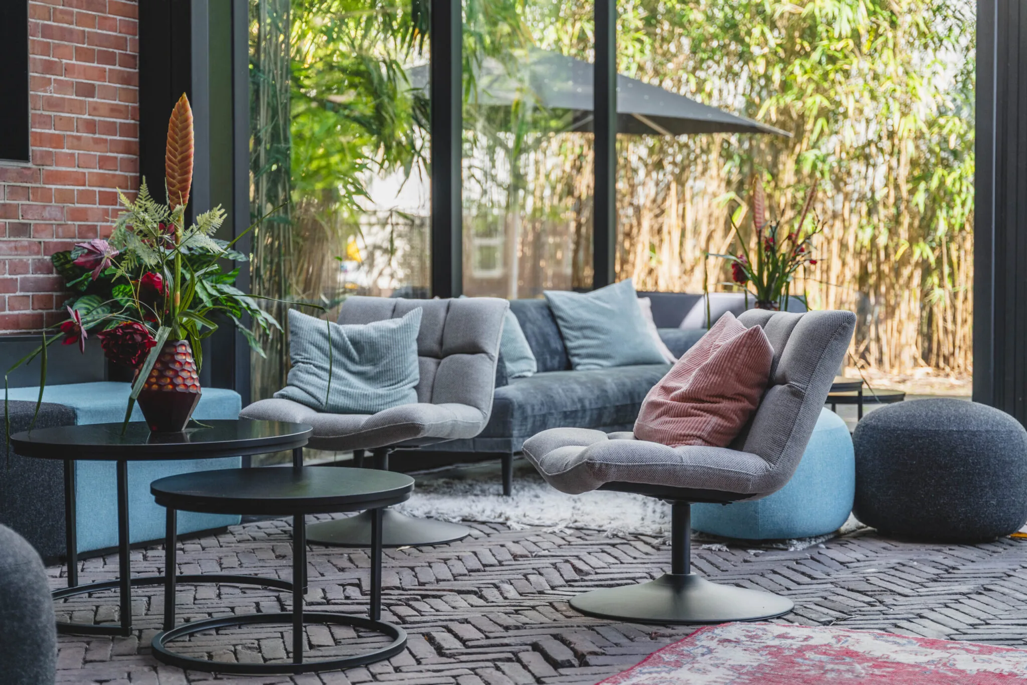 Stijlvol ingerichte loungehoek op Raamplein 1-9 met moderne meubels, kussens, planten en grote ramen met uitzicht op bamboe.