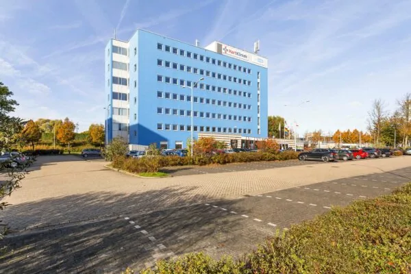 Een helderblauw kantoorgebouw genaamd Ravelijn 1 met parkeervoorziening op de voorgrond, omringd door herfstkleurige bomen.