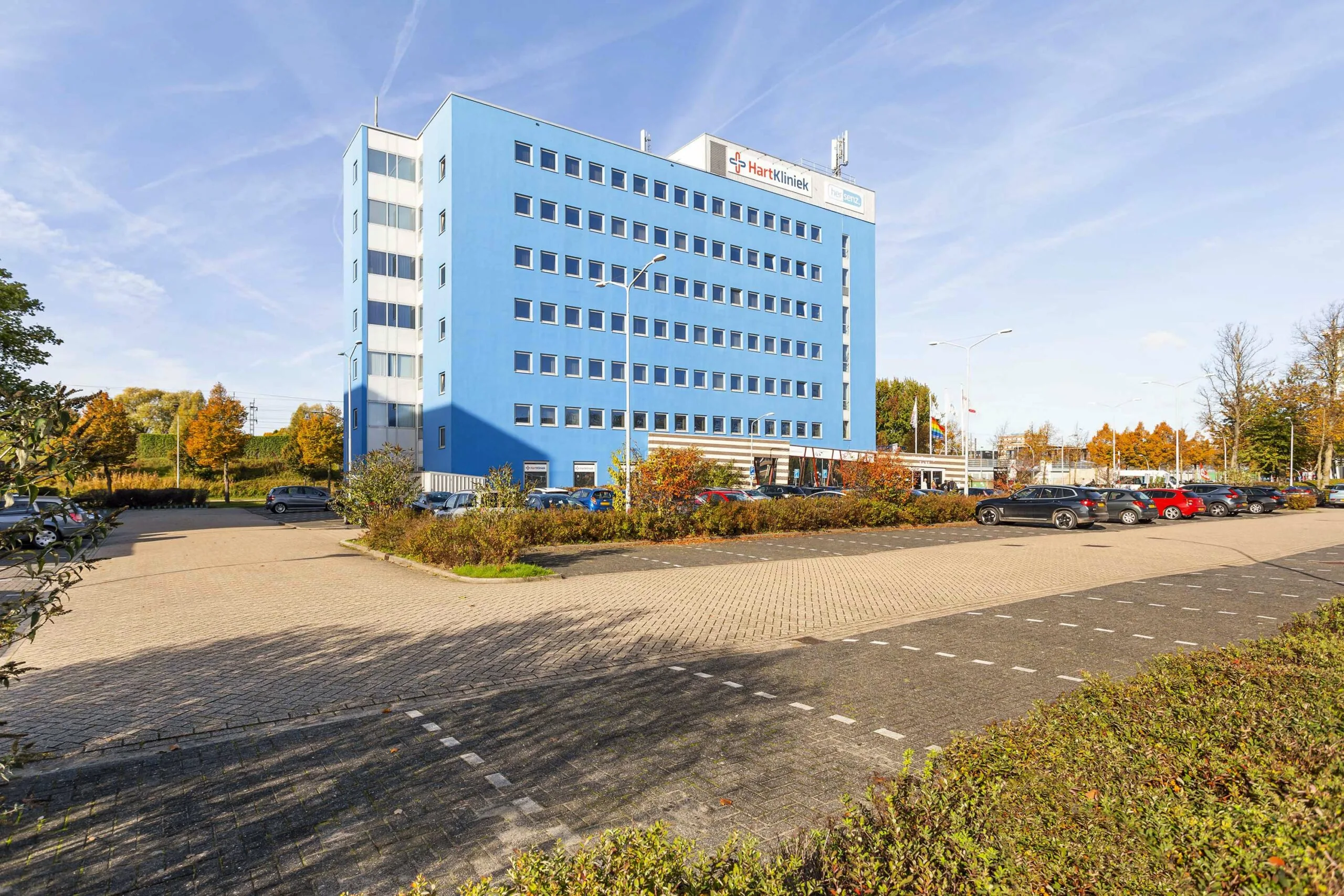 Een helderblauw kantoorgebouw genaamd Ravelijn 1 met parkeervoorziening op de voorgrond, omringd door herfstkleurige bomen.
