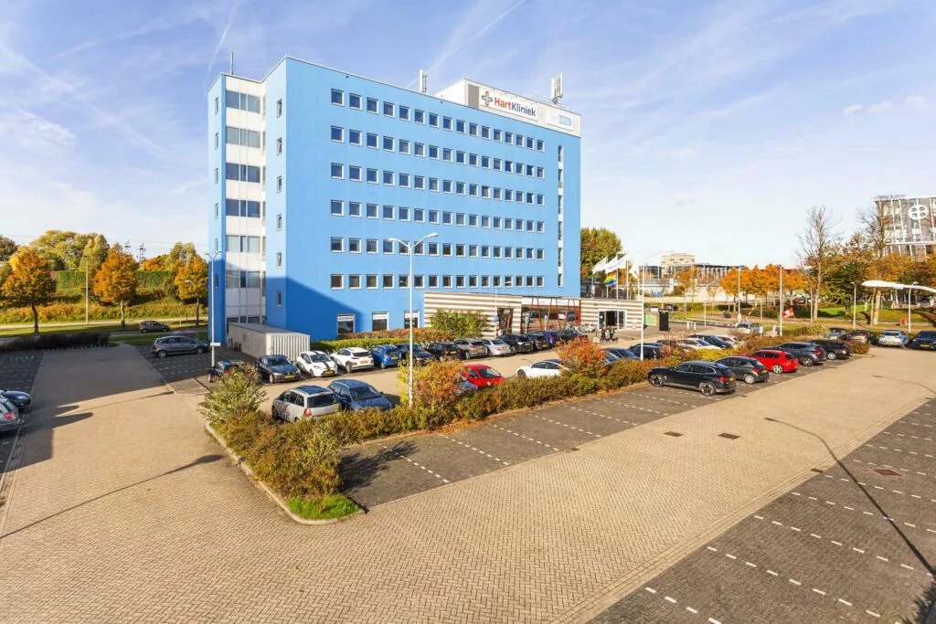 Blauw kantoorgebouw Ravelijn 1 met HartKliniek en parkeerplaats op een zonnige herfstdag.