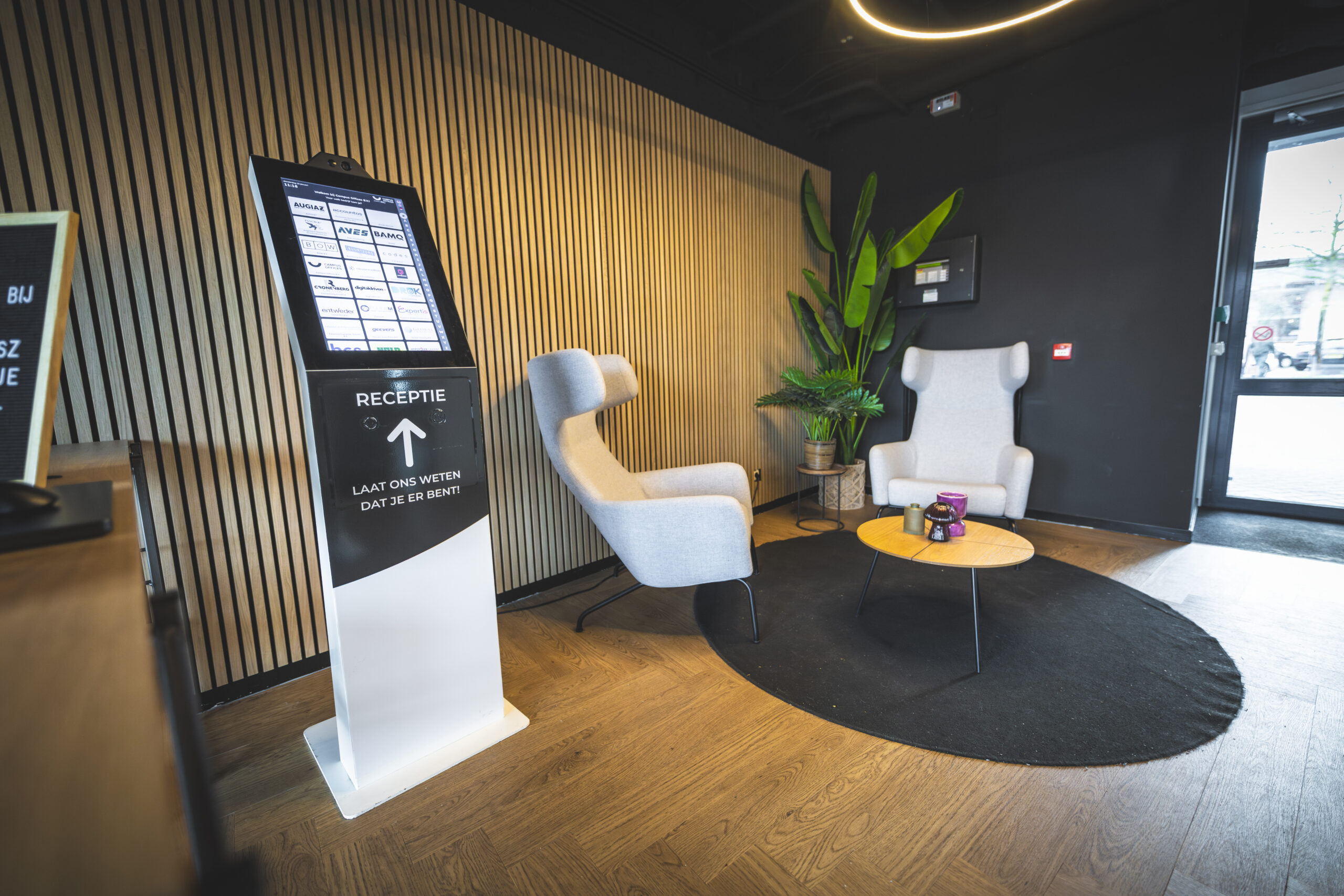 Moderne receptieruimte met twee grijze stoelen, een touchscreen aanmeldzuil en een ronde tafel op een zwart vloerkleed.