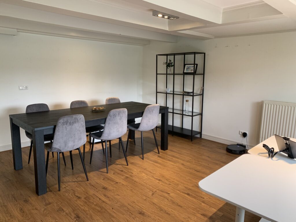 Vergaderruimte met een zwarte tafel, zes grijze stoelen, een open vakkenkast en een wit bureau.