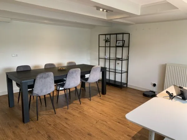 Vergaderruimte met een zwarte tafel, zes grijze stoelen, een open vakkenkast en een wit bureau.