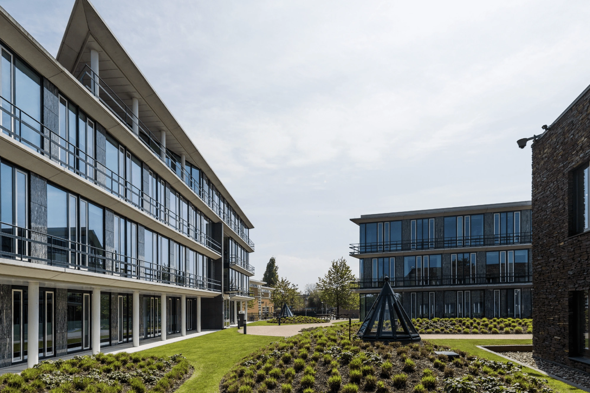 Moderne kantoorgebouwen met veel glas rondom een groen aangelegde binnentuin met kunstobjecten.