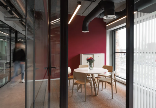 Een vergaderruimte met een ronde tafel, vier stoelen en een vaas met bloemen, tegen een bordeauxrode muur.