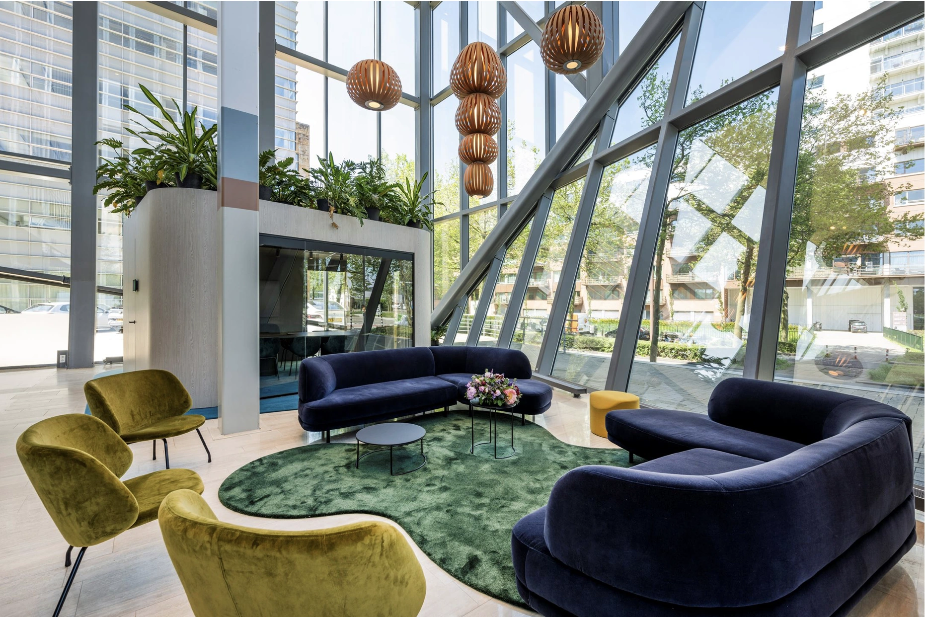 Moderne en lichte lobby met grote ramen, fluwelen zitmeubels, groene planten en houten hanglampen.