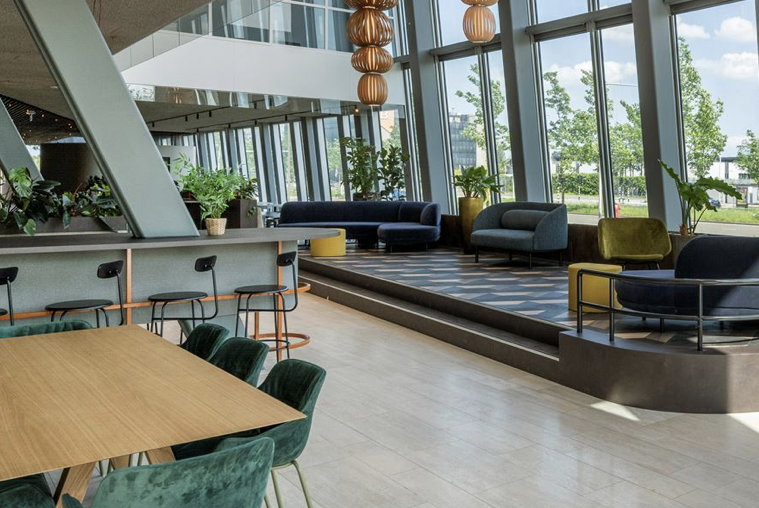 Moderne en lichte kantoorlounge met zithoeken, hoge ramen en groenaccenten.