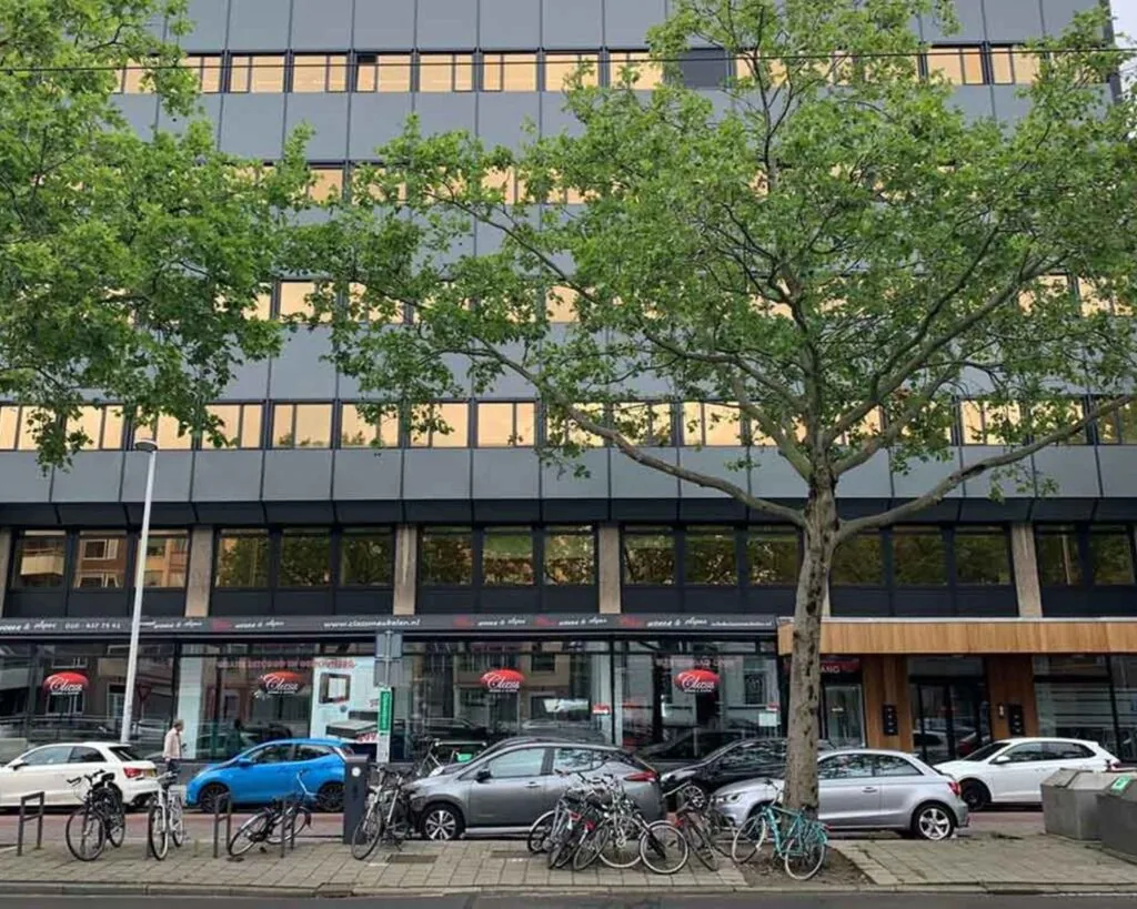 Vooraanzicht van het kantoorgebouw aan de Schiekade 34 in Rotterdam met geparkeerde auto’s, fietsen en bomen op de stoep.