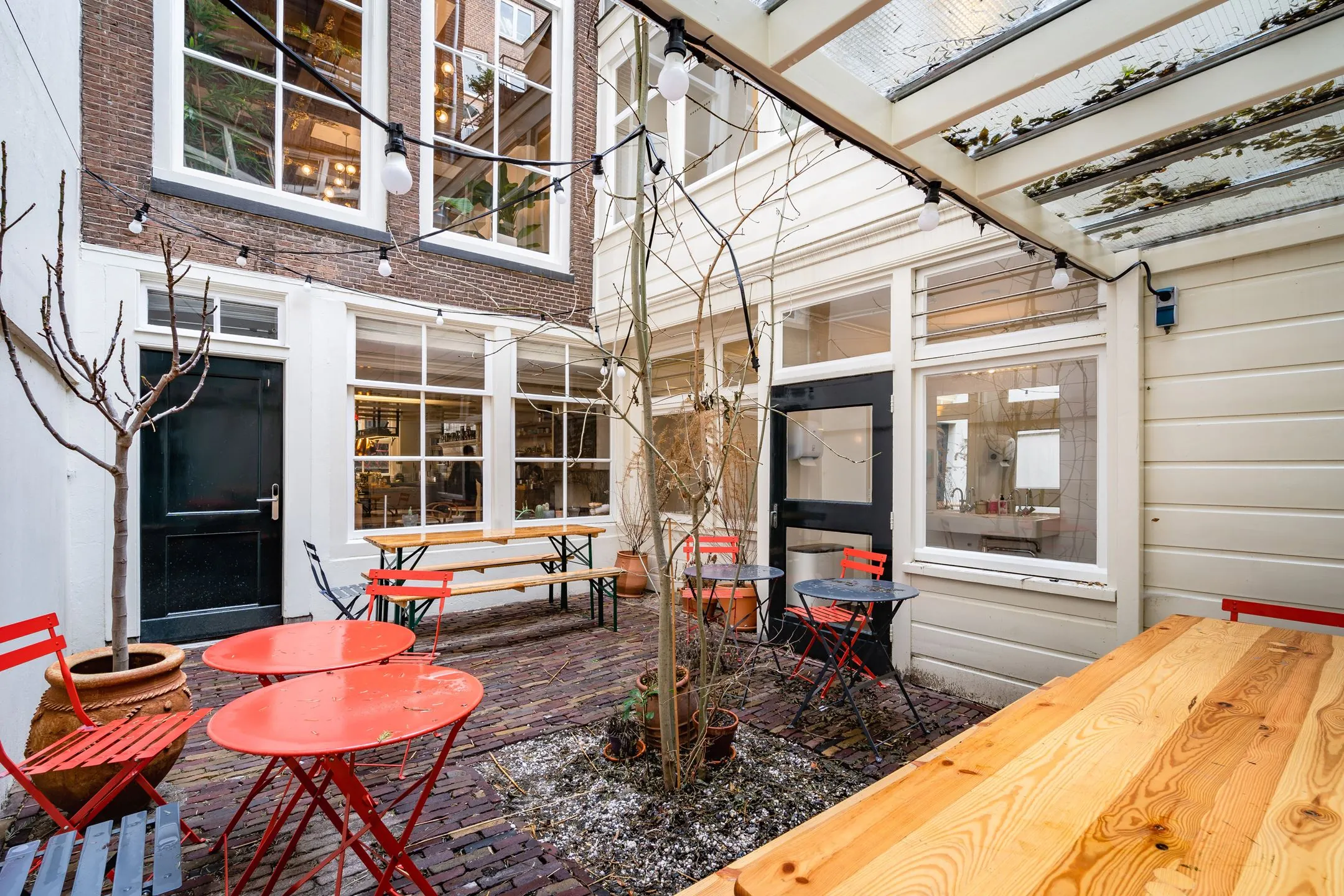 Knusse binnentuin van Singel 258_9 met rode bistrostoelen, houten tafels en sfeerverlichting onder een gedeeltelijk glazen dak.