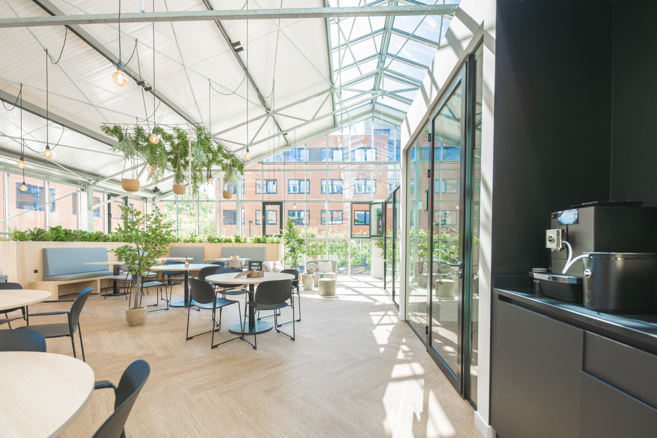 Lichte en moderne ontmoetingsruimte "Sociaal hart 10 groot" met veel planten, zitplekken en glazen dak.