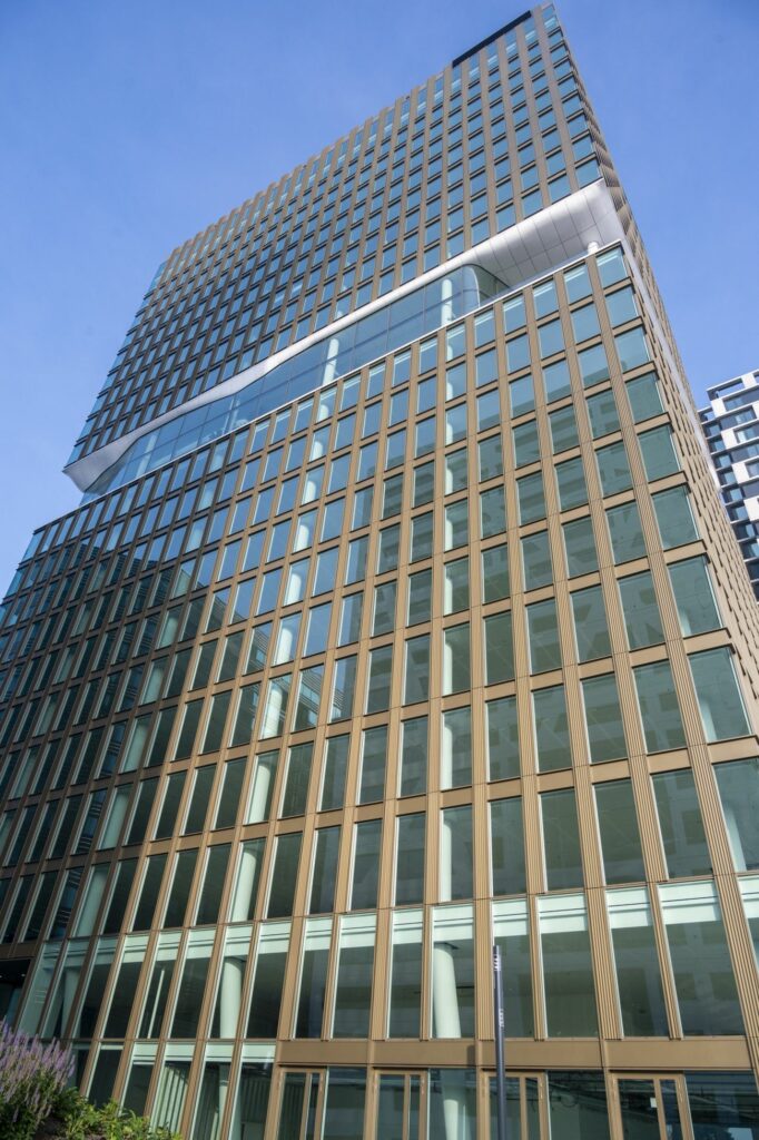 Hoog gebouw met glazen gevel en bronzen raamkozijnen aan het Stadsplateau 27-29 in Utrecht, gefotografeerd vanaf straatniveau tegen een blauwe lucht.