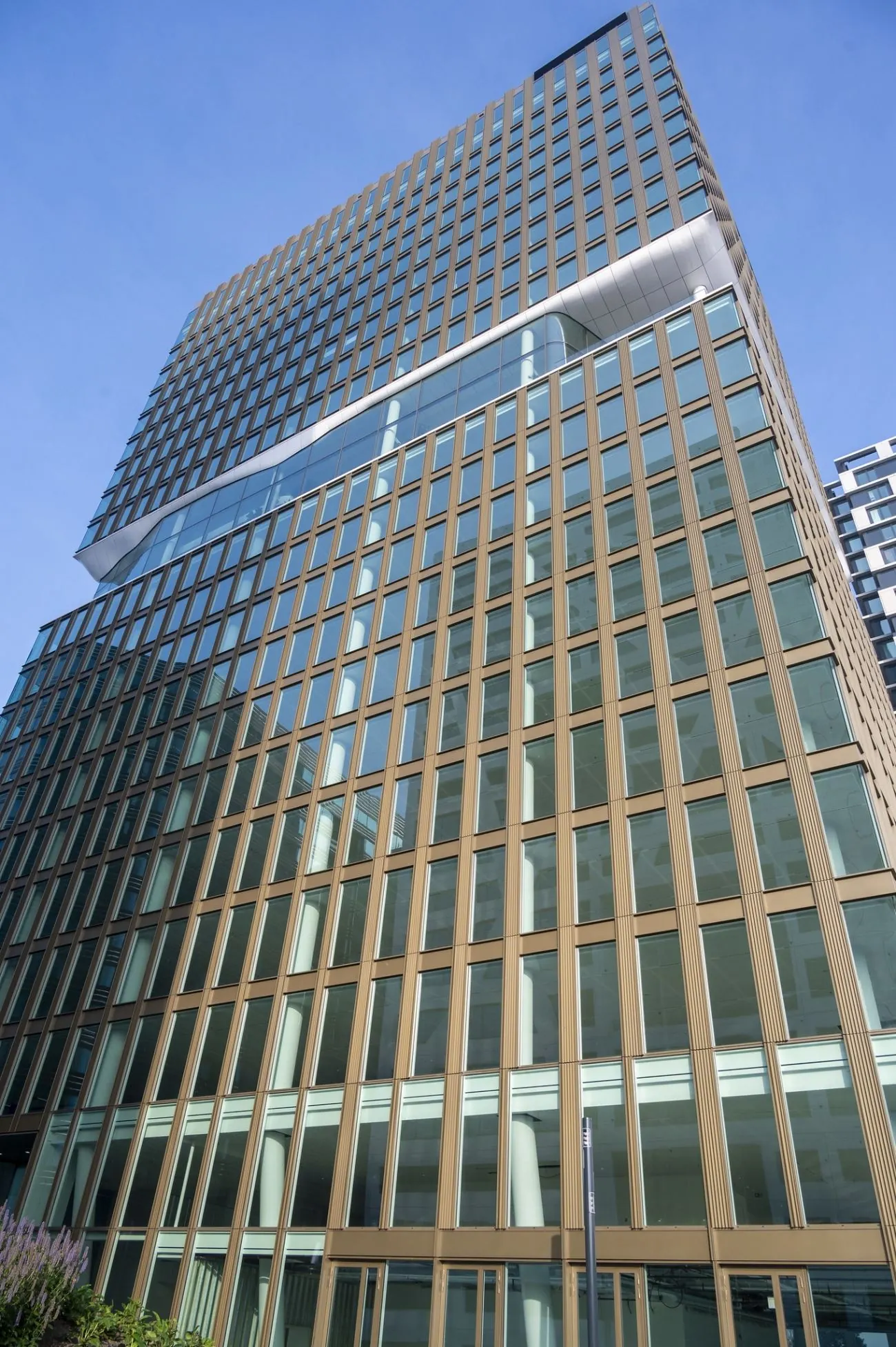 Hoog gebouw met glazen gevel en bronzen raamkozijnen aan het Stadsplateau 27-29 in Utrecht, gefotografeerd vanaf straatniveau tegen een blauwe lucht.
