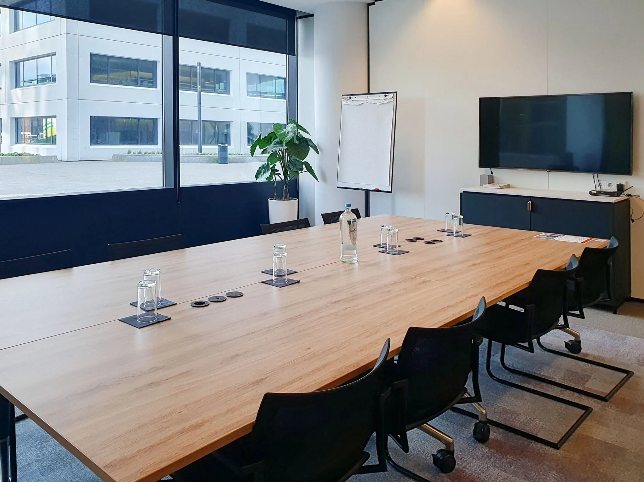 Vergaderruimte in Stadsplateau 7 met grote houten tafel, stoelen, flip-over, televisiescherm en uitzicht op modern kantoorgebouw.