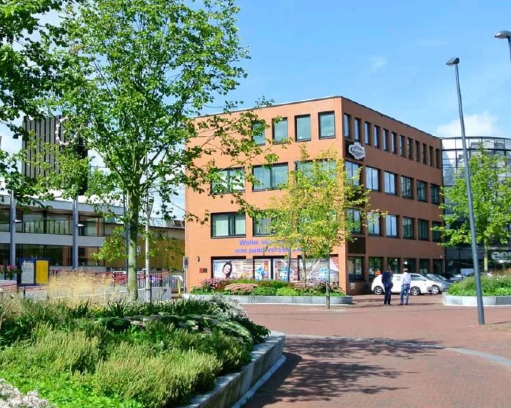Een modern kantoorgebouw aan het Stationsplein met omliggende bomen, planten en enkele passerende voetgangers.