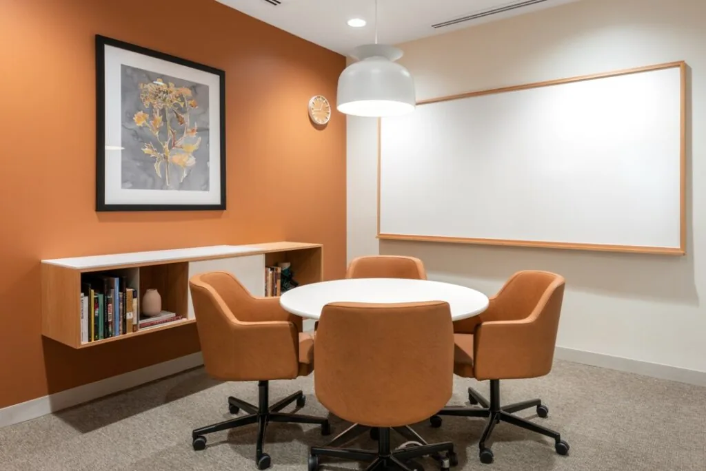 Kleine vergaderruimte met ronde tafel, vier oranje stoelen, een whiteboard en een wandkast met boeken.