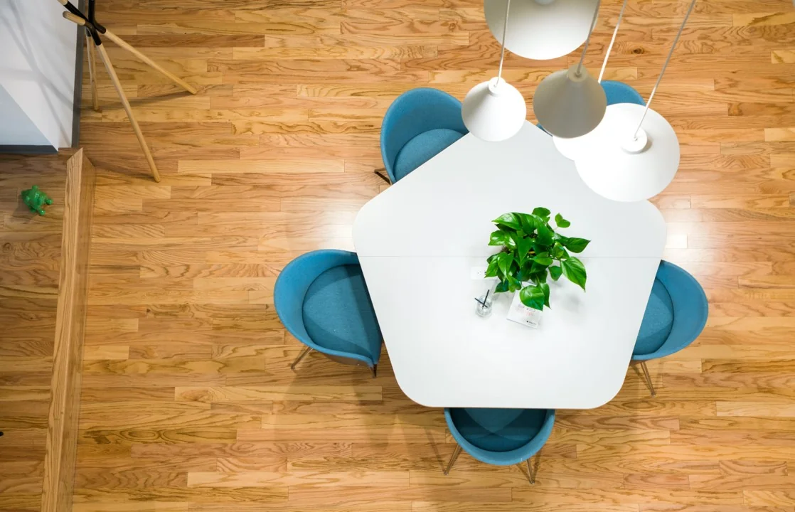 Wit vierkant tafelblad met plant en vier blauwe stoelen op houten vloer, gezien van bovenaf.