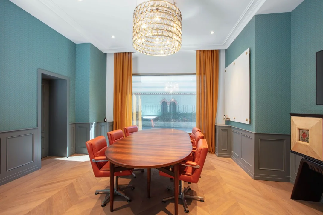 Vergaderruimte met ovaal houten tafel, roodbruine stoelen, grote kroonluchter en oranje gordijnen in Stationsplein 18.