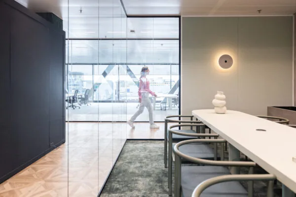 Modern kantoorinterieur van The Joan-11 met vergadertafel op de voorgrond en een persoon die achter glazen wanden voorbijloopt.