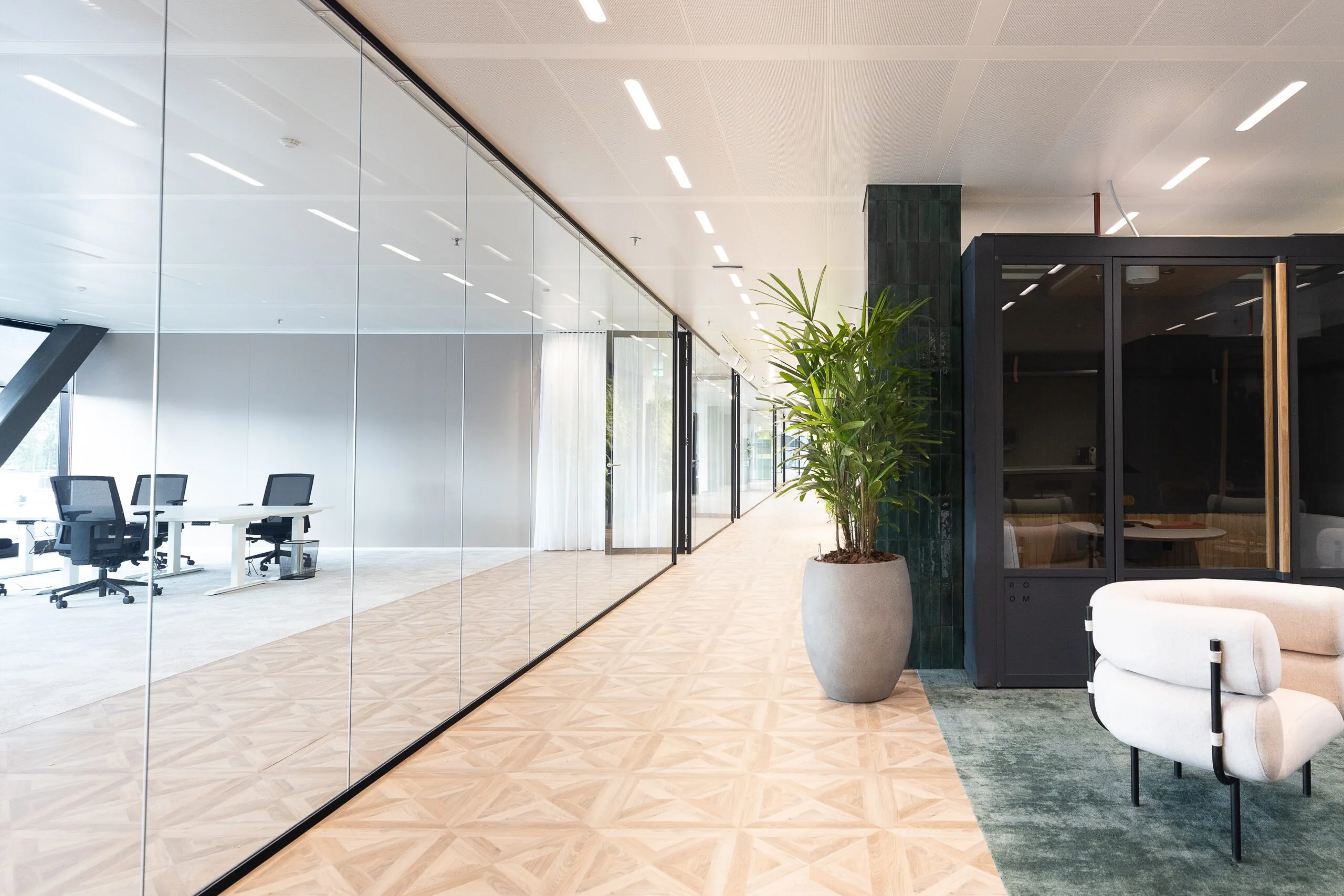 Modern kantoorinterieur van The Joan-13 met glazen vergaderruimtes, designmeubilair en een grote plant in een strakke gang.