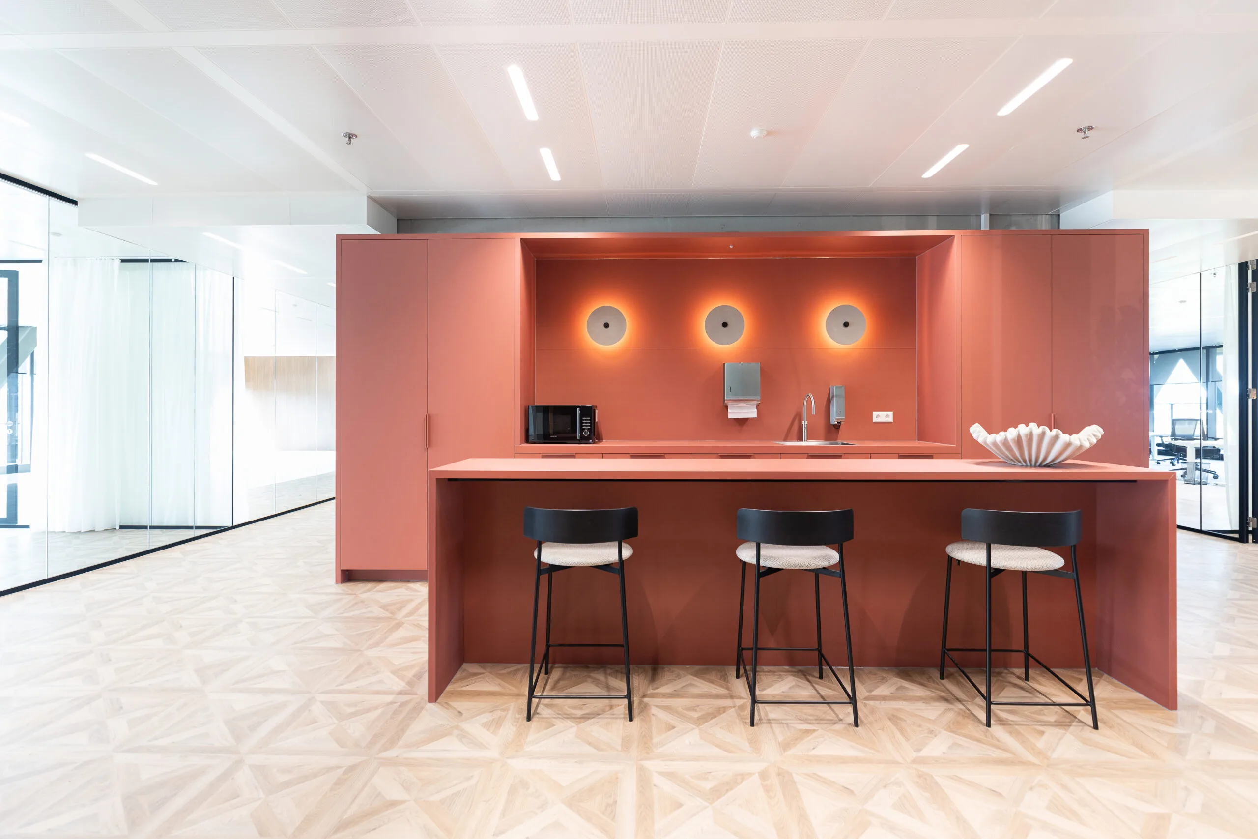 Moderne kantoorkeuken in The Joan-19 met terracottakleurig keukeneiland, drie barkrukken en minimalistisch design.