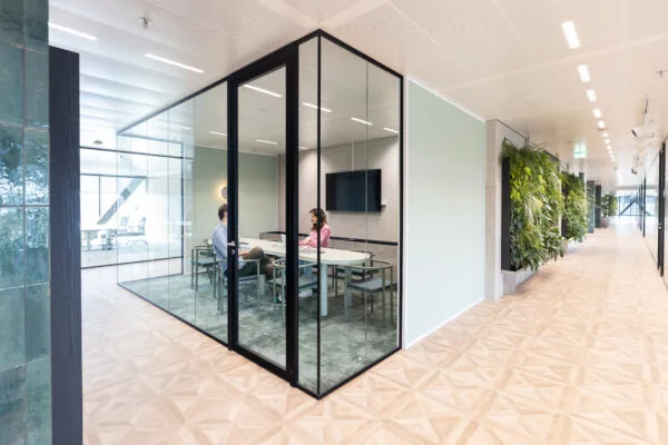 Twee mensen voeren een gesprek in vergaderruimte The Joan-9 in een modern en licht kantoorpand met glazen wanden en een groene plantenwand in de gang.