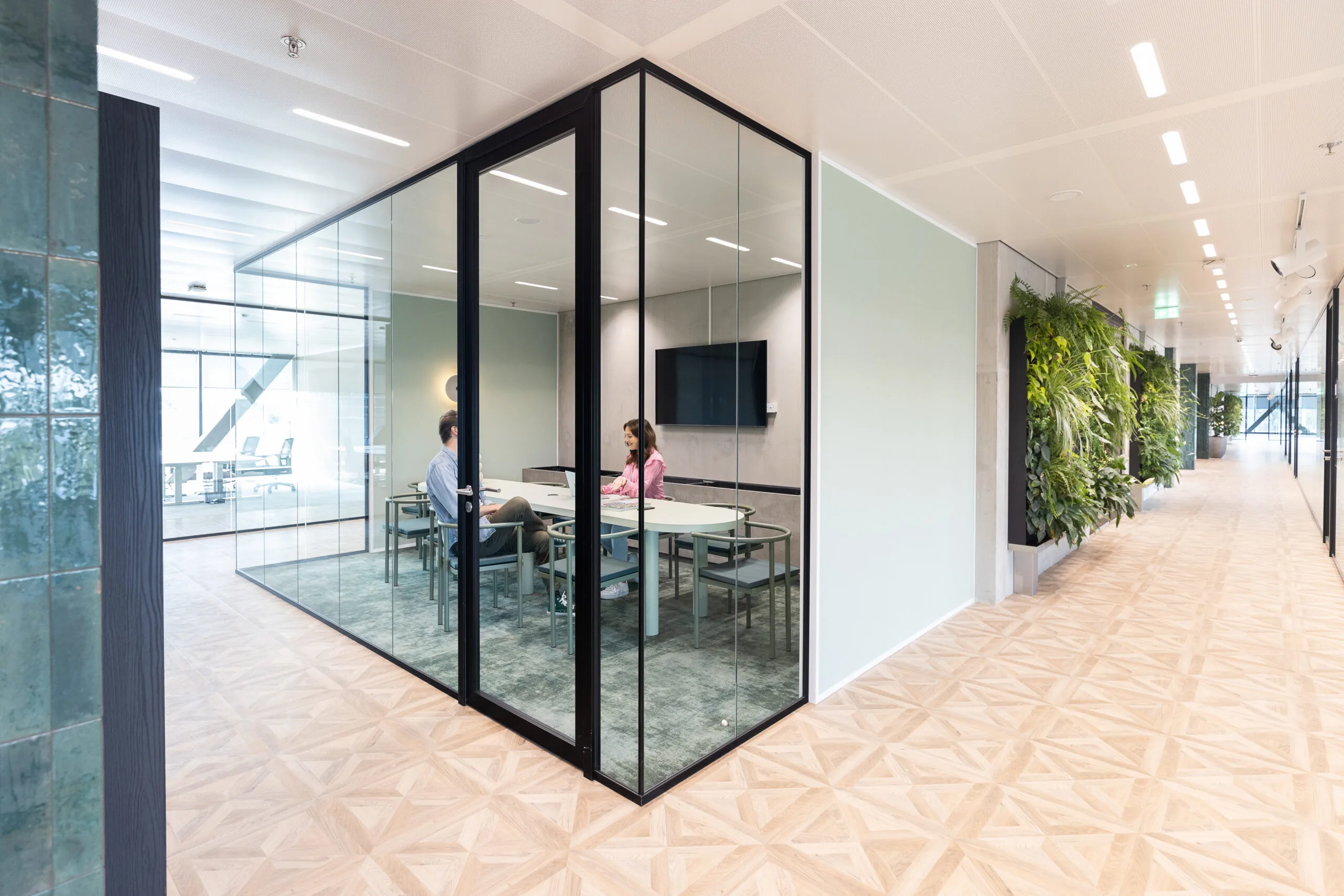 Twee mensen voeren een gesprek in vergaderruimte The Joan-9 in een modern en licht kantoorpand met glazen wanden en een groene plantenwand in de gang.