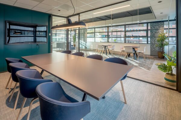 Moderne vergaderruimte aan de Tielweg 30 met glazen wand, grote tafel, blauwe stoelen en zicht op een lunchruimte.